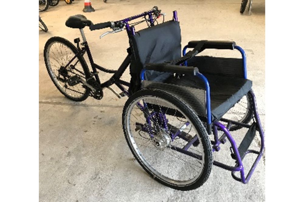 A wheelchair bike with a wheelchair frame on the front for one rider to sit in. The bike is powered by a second rider on a saddle pedalling at the back