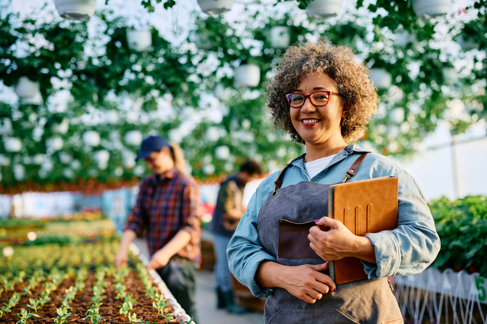 Smiling woman in a greenhouse holding a notebook beside rows of young plants while others work in the background.