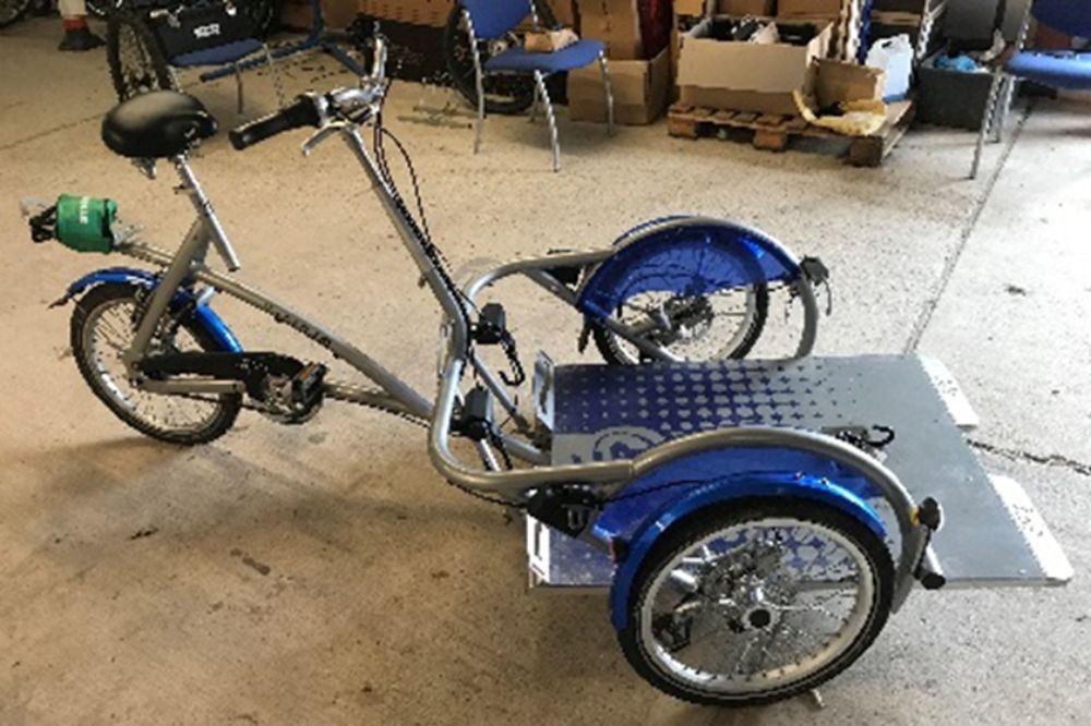 A platform for a wheelchair is at the front of the Vanraam wheelchair bike. The bike is powered by a second rider who sits on the saddle behind the wheelchair platform and pedals