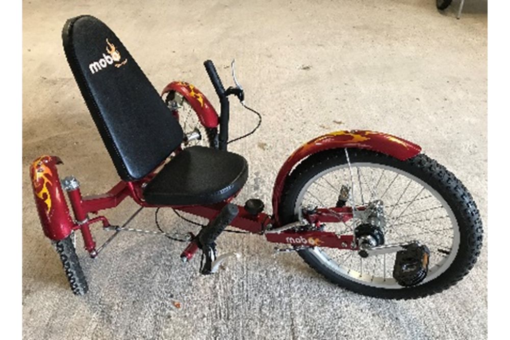 A red recumbent trike. The rider sits in the seat leaning against the backrest and uses the pedals on the front wheel to power the bike. The handlebars are either side of the seat