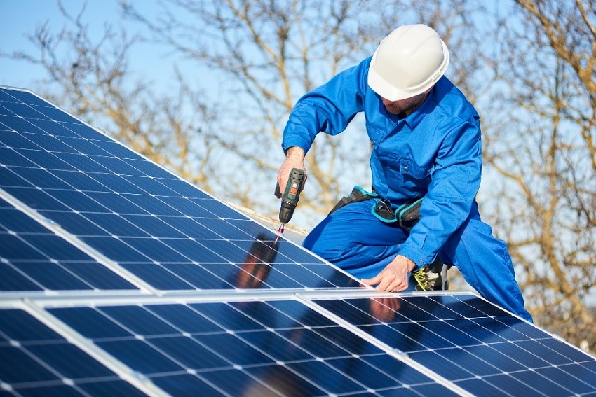 A man in blue overalls and a hard hat installs solar panels using a drill