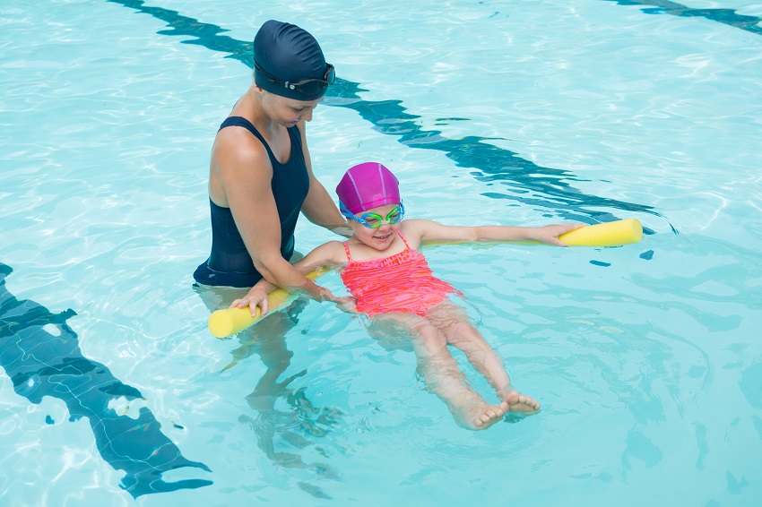 A swimming instructor helps a girl to swim on her back with a yellow pool noodle