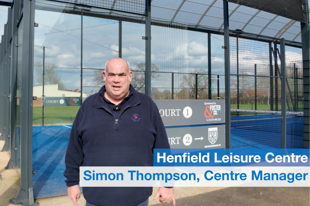 A man identified on-screen as Simon Thompson, Centre Manager, stands in front of fenced outdoor courts at Henfield Leisure Centre, speaking to camera on a bright day.
